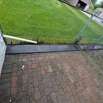 Paved walkway leading to a fenced yard with green grass and a drainage pipe.