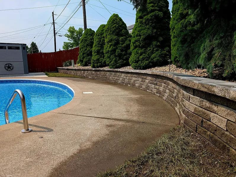 Curved stone wall surrounding a blue swimming pool with lush greenery and clear sky.
